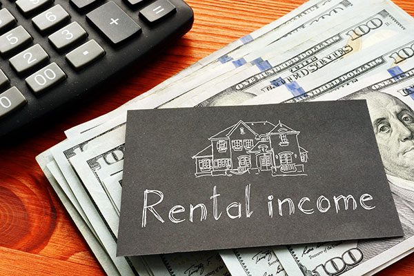 Black business card with a drawing of a house and text that says, "rental income" sits on top of a stack of $100 bills next to a calculator on a wooden desk